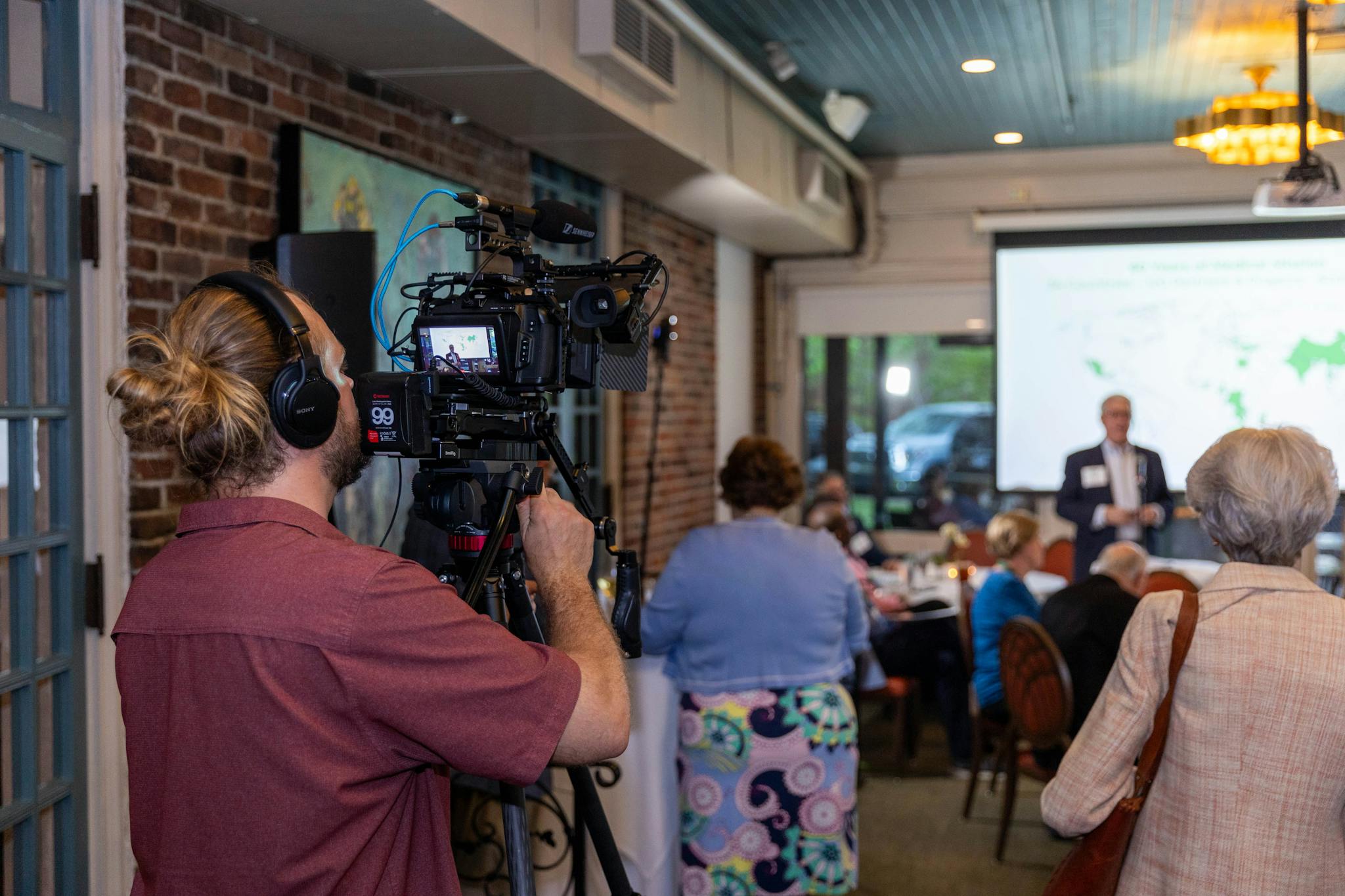 Indoor scene of a videographer documenting a professional corporate event with attendees and a projection screen.